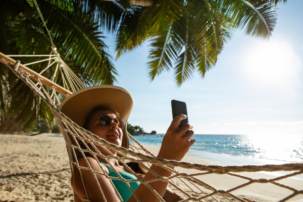 A person using a smartphone with E-SIM while relaxing in a beach hammock under palm trees