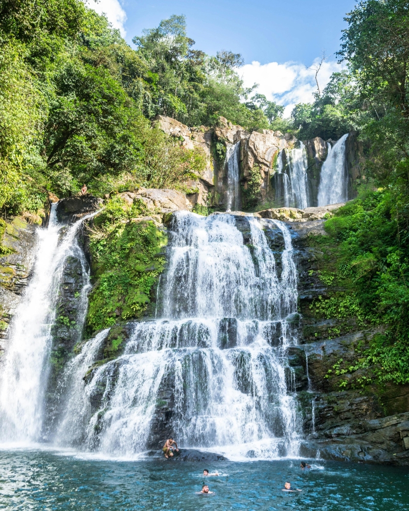 Large cascading waterfall with multiple tiers and swimmers enjoying natural pool in tropical setting