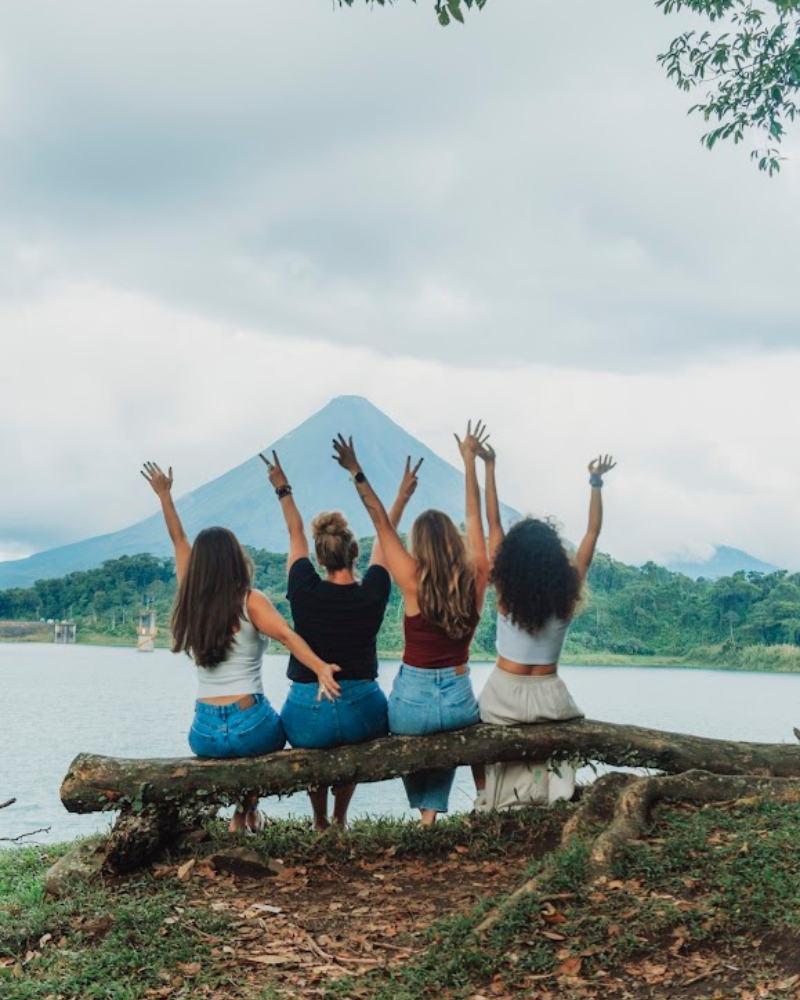 Four friends sitting on log overlooking Lake Arenal and volcano, raising hands in celebration